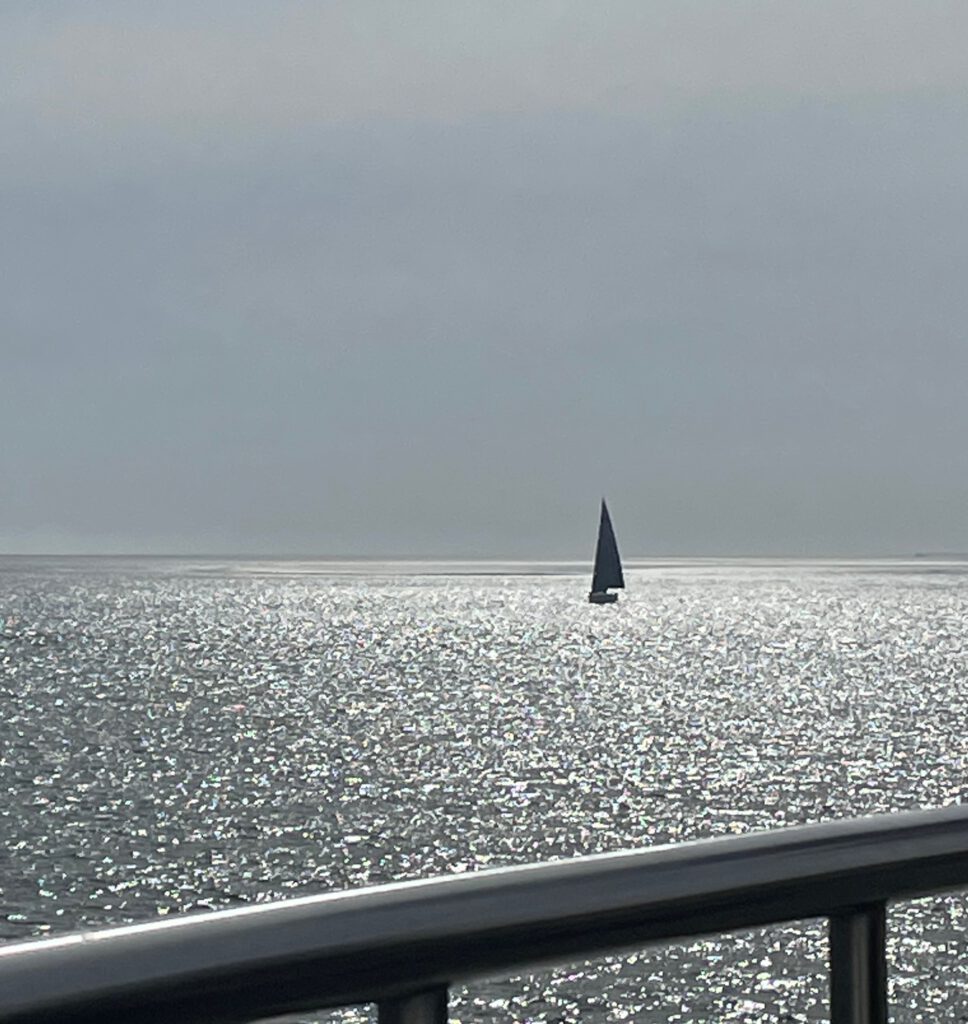 Ein kleines Segelboot hebt sich am Horizont dunklgrau vor grauem Himmel ab. Das Meer glitzert silbrig. Am unteren Bildrand sieht man Teile einer Reling.